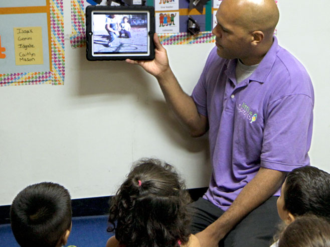 A teacher shows a video on an iPad, which he holds up in front of a group of students.