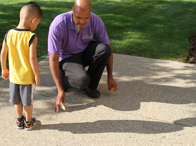 A student stands on the sidewalk and looks at his shadow. A teacher crouches next to him with a piece of chalk in his hand.