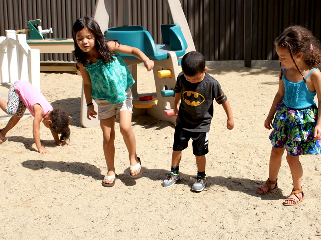 Four students are playing in a sandy playground. It is sunny, and their shadows are visible.