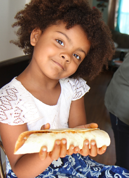 A little girl holds a rolled tortilla and smiles.