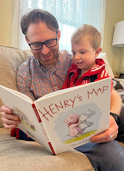 A father reads the book “Henry’s Map” by David Elliot to a child.