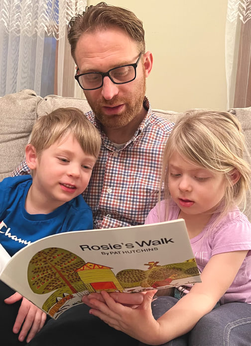 A father and his two children read the book “Rosie’s Walk” by Pat Hutchins.