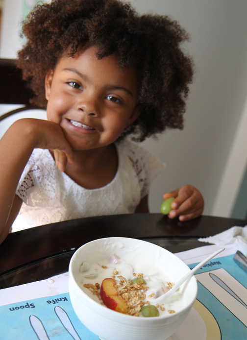 A little girl smiles and holds a grape while she sits at a table with a bowl of yogurt with fruit and granola.