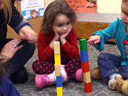 A preschool girl in a blue shirt points to one of two block towers. A child in a red shirt and the teacher watch her count.