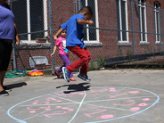 A preschool boy in a blue and red outfit jumps above a chalk circle with lines dividing it into slices. A teacher and another child watch.