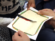 Close-up of a child cutting a paper sandwich held by a teacher.