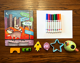 Julie Dillemuth’s book “Lucy in the City” next to a stack of paper, markers, and small, differently shaped objects laid out on a table.