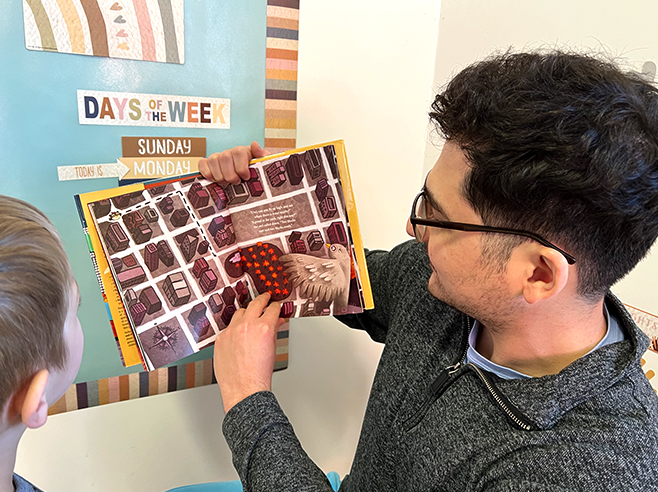 A man points to an aerial view of a map in the book “Lucy in the City” by Julie Dillemuth while a child watches.