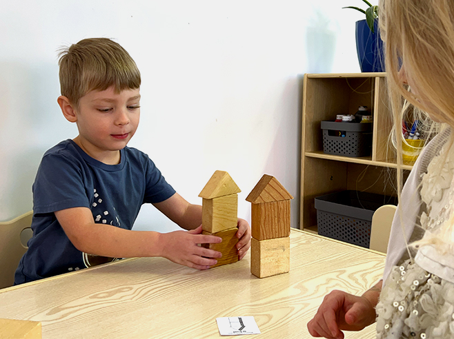 A boy and a girl position two different block towers in relation to each other based on a paper card on a table.