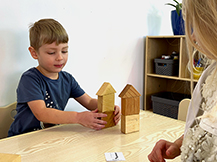 A boy and a girl position two different block towers in relation to each other based on a paper card on a table.