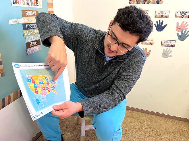 A man sits on a chair and points to a map of the United States of America.