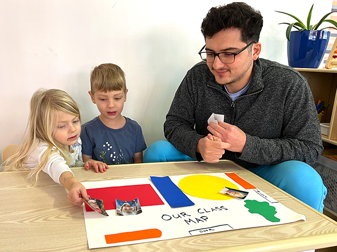 A man sits with a boy and a girl at a table while they place pictures of children on a handmade map of a classroom.