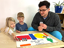 A man sits with a boy and a girl at a table while they place pictures of children on a handmade map of a classroom.