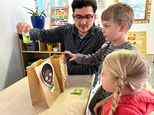 A man holds up a card showing a spatial relationship between a cartoon girl and a goat while two kids position paper bags with the characters.