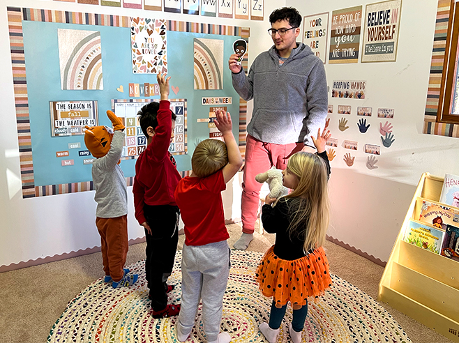 A man holds up a cartoon character cutout of Gracie while children hold up their right hands in a preschool classroom.