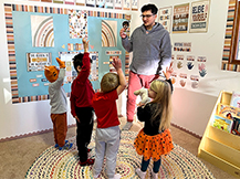 A man holds up a cartoon character cutout of Gracie while children hold up their right hands in a preschool classroom.