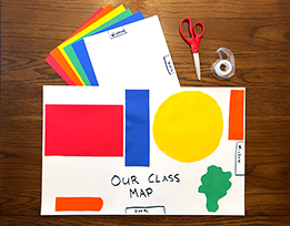 A handmade map of a classroom with objects depicted by brightly colored shapes, next to scissors, tape, and a stack of paper.