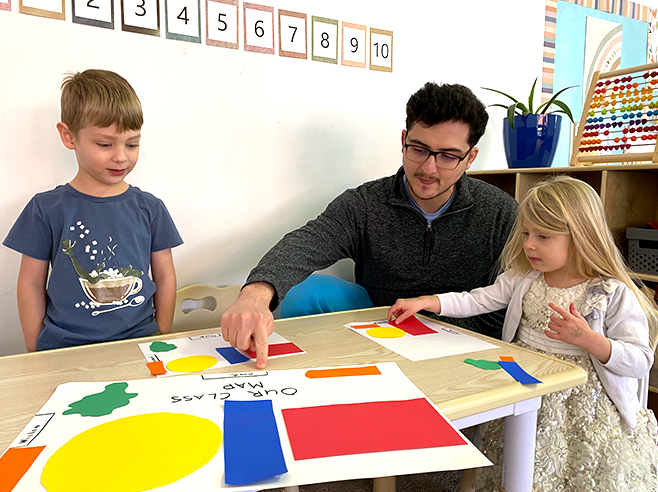 A man sits at a table and arranges brightly colored shapes onto a piece of blank paper while a girl and a boy watch.