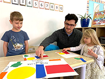 A man sits at a table and arranges brightly colored shapes onto a piece of blank paper while a girl and a boy watch.