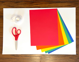 A stack of colored paper next to scissors and tape on top of a large white sheet of cardstock.