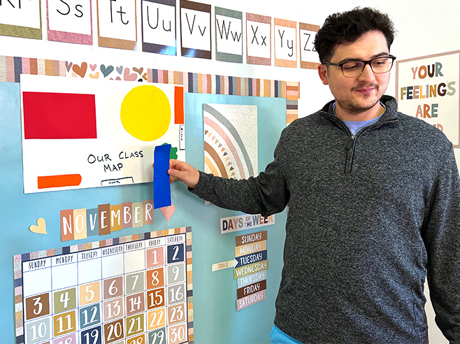 A man stands in front of a preschool bulletin board and places a blue rectangle on a handmade map of the classroom.