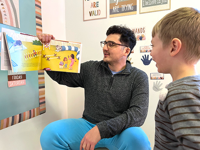 A man holds up and reads Julie Dillemuth’s book “Mapping My Day” while a child watches and smiles.