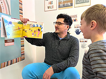 A man holds up and reads Julie Dillemuth’s book “Mapping My Day” while a child watches and smiles.
