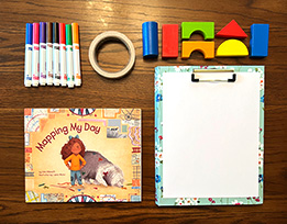 Julie Dillemuth’s book “Mapping My Day” next to a stack of paper on a clipboard, a pile of markers, tape, and differently-shaped blocks.