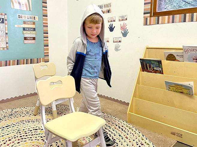 A boy walks between two chairs in a preschool classroom.