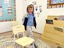 A boy walks between two chairs in a preschool classroom.