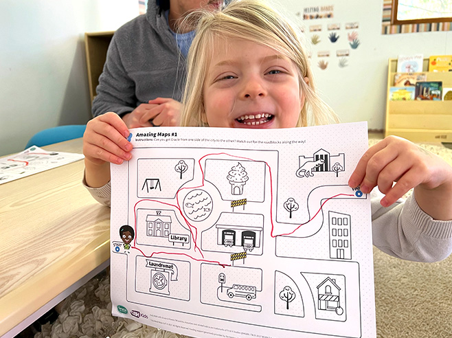 A smiling girl holds up a printout of a maze with a pathway hand drawn from the start to finish.