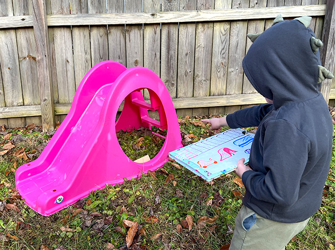 A boy holds a clipboard with a treasure map while he points to a small box under a slide.