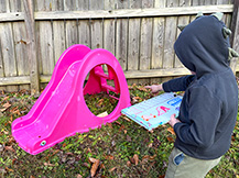 A boy holds a clipboard with a treasure map while he points to a small box under a slide.