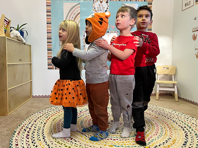 Four children stand in a line in the classroom holding onto the shoulders of the child in front.