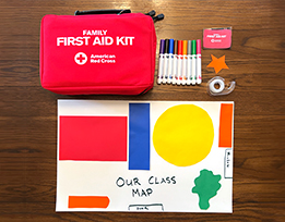 A handmade map of a classroom, a red first aid kit, a picture of a first aid kit, markers, tape, and a paper star laid out on a table.