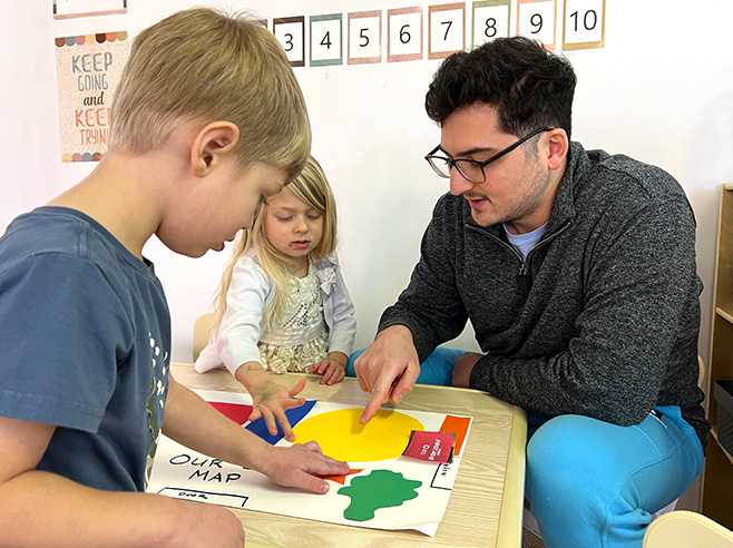 A man, a boy, and a girl point to colorful shapes that represent objects on a large handmade map of a classroom.