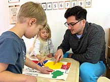 A man, a boy, and a girl point to colorful shapes that represent objects on a large handmade map of a classroom.