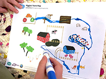 A boy draws a pathway with a marker between, over, around, and through landmarks on a printout of a farm map.