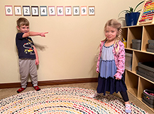 A boy points to a girl who is standing and smiling in a preschool classroom.