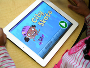 A preschool girl in a pink striped shirt looks at the Gracie and Friends City Skate app on an iPad.