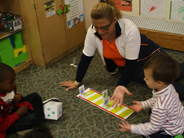 A preschool teacher and two children play the Jungle Gym Board Game on the carpeted floor.