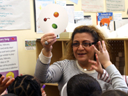 Preschoolers hold up three fingers as their teacher shows a round card with pictures of three fruits on it.