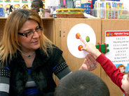 A young child points to a picture of an orange on a round paper card held by her teacher.