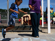 A boy hops over a chalk square drawn on pavement with dots in it. A teacher stands nearby.