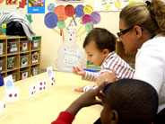 A teacher watches as a child reaches out to place a card in one of five cups with different numbers of dots.