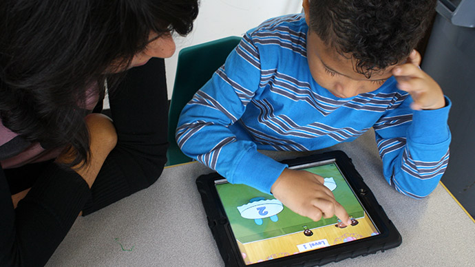 A preschool boy in a blue striped shirt plays the Early Math with Gracie & Friends Birthday Cafe app and drags cartoon children to a table while a teacher in a black sweater sits beside him watching.