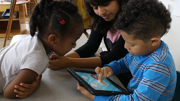 A preschool boy in a blue striped shirt plays the Early Math with Gracie & Friends Breakfast Time app and swipes to cut a waffle while a preschool girl and a teacher sit nearby and look on.