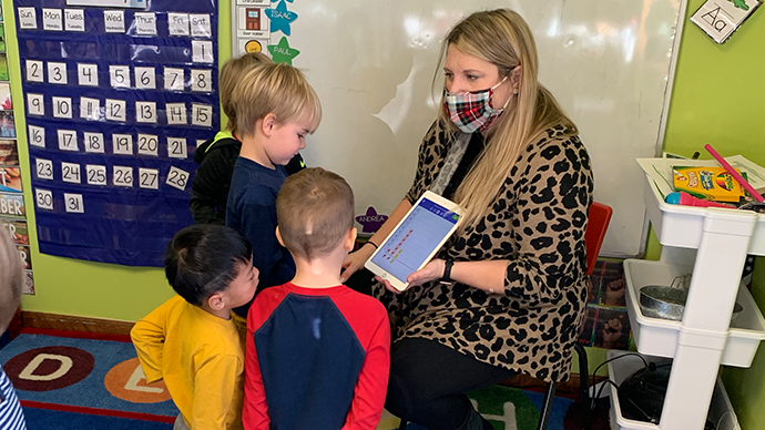 A teacher holds up an iPad displaying a graph while children gather close together looking at the screen.