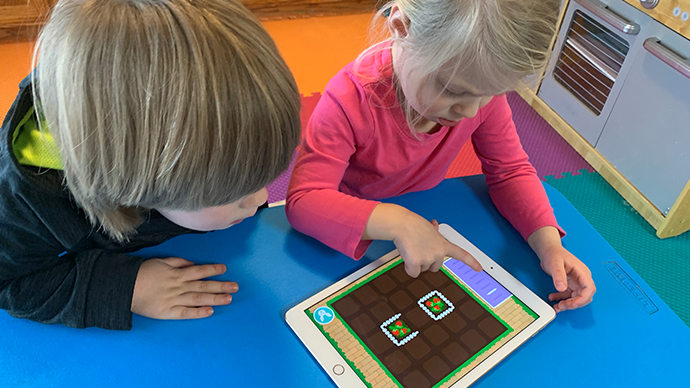 Two children are sitting at a blue table and playing an iPad displaying the Berry Garden app, which has a brown field with two patches of berries planted.  