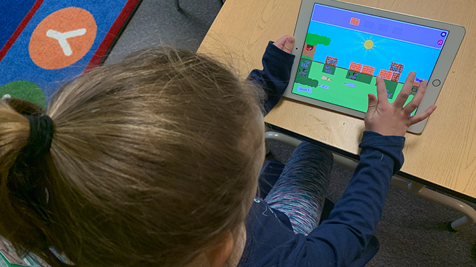 A child is sitting at a laminated wood table and playing an iPad displaying the Puppy Park app, which has a sunny sky and blocks casting square shadows onto grass. 